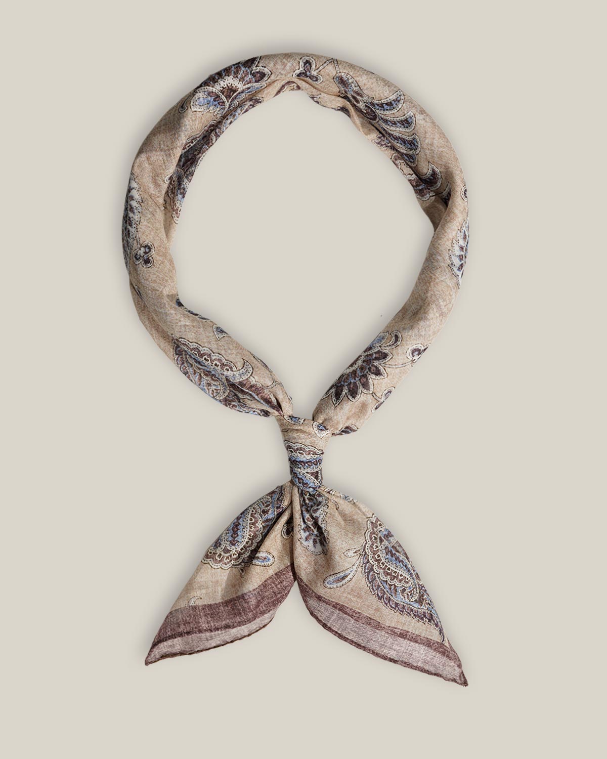 Beige cotton bandana with intricate dark paisley motifs and a muted contrasting border, tied as a neck scarf.