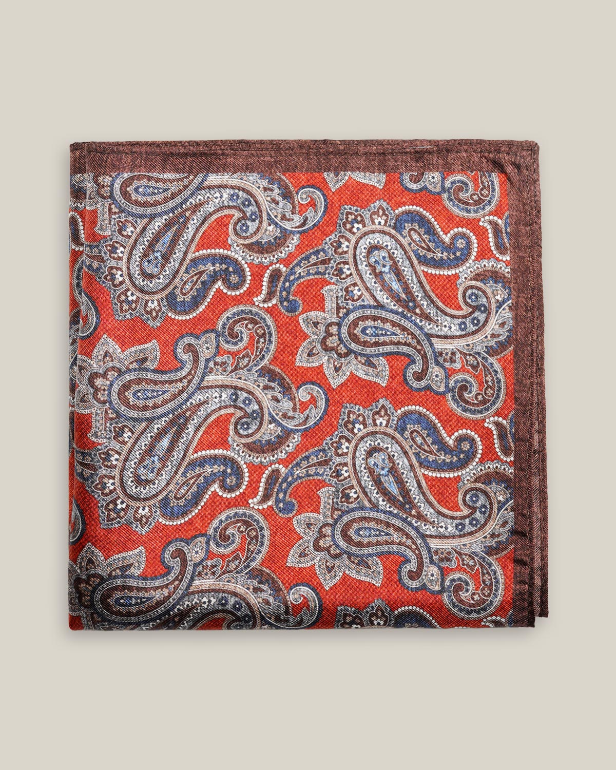Rust-red printed silk pocket square with large brown, ivory and muted blue paisley motifs and a contrasting brown border.