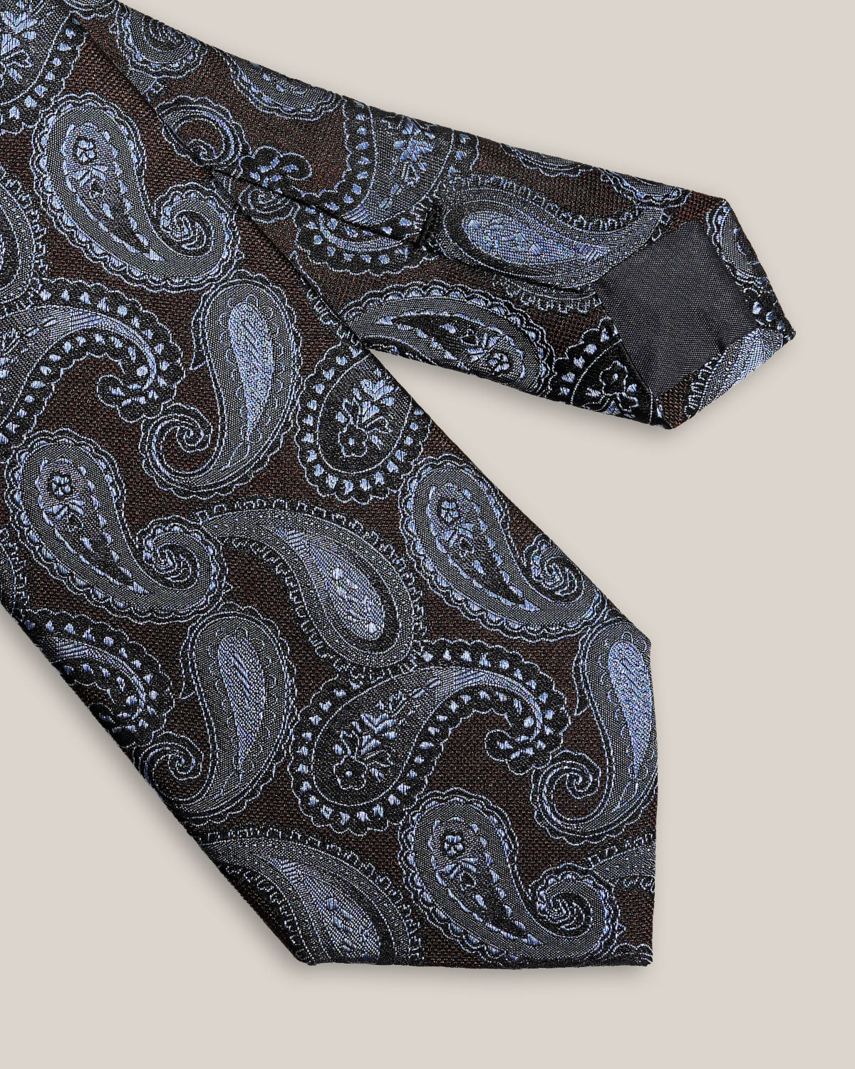 Close-up of light blue and silver paisley motifs on a dark brown 100% silk tie.